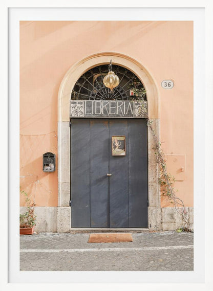 A charming European bookstore entrance featuring a large, dark grey arched door set against a peach-colored wall. Above the door, a sign reads 'LIBRERIA' below a decorative fanlight window and a hanging lantern. Decor