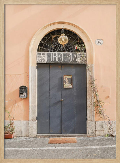 A charming European bookstore entrance featuring a large, dark grey arched door set against a peach-colored wall. Above the door, a sign reads 'LIBRERIA' below a decorative fanlight window and a hanging lantern. Decor