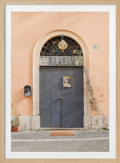 A charming European bookstore entrance featuring a large, dark grey arched door set against a peach-colored wall. Above the door, a sign reads 'LIBRERIA' below a decorative fanlight window and a hanging lantern. Decor