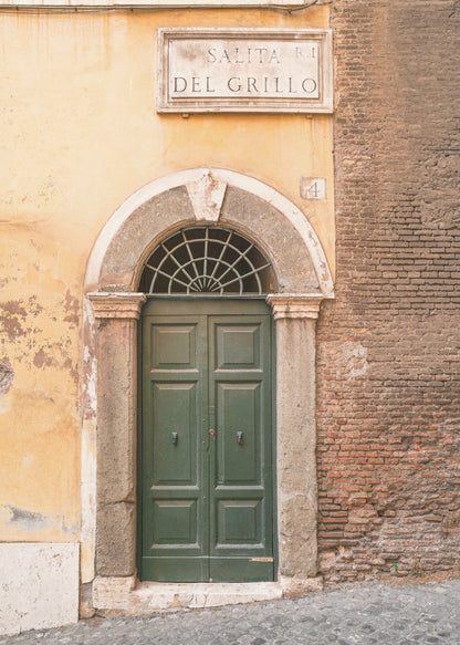 A vertical shot of a rustic European street scene featuring a dark green arched wooden door set into a stone frame. The building's facade is a mix of peeling yellow ochre plaster and exposed red brick. Above the door is a marble plaque that reads 'SALITA RI DEL GRILLO', and the ground is paved with grey cobblestones. Wall Art