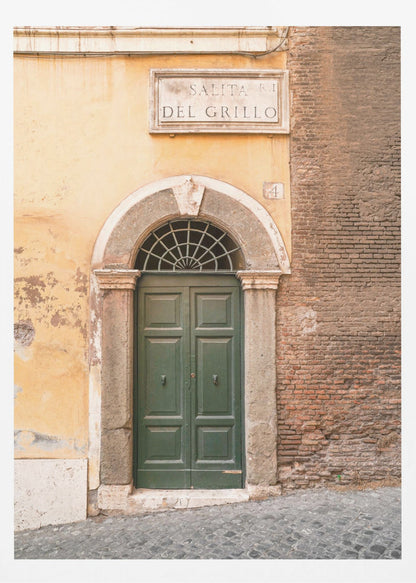 A vertical shot of a rustic European street scene featuring a dark green arched wooden door set into a stone frame. The building's facade is a mix of peeling yellow ochre plaster and exposed red brick. Above the door is a marble plaque that reads 'SALITA RI DEL GRILLO', and the ground is paved with grey cobblestones. Wall Art