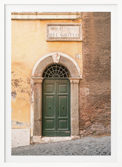 A vertical shot of a rustic European street scene featuring a dark green arched wooden door set into a stone frame. The building's facade is a mix of peeling yellow ochre plaster and exposed red brick. Above the door is a marble plaque that reads 'SALITA RI DEL GRILLO', and the ground is paved with grey cobblestones. Wall Art