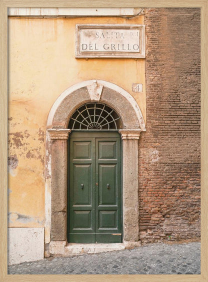 A vertical shot of a rustic European street scene featuring a dark green arched wooden door set into a stone frame. The building's facade is a mix of peeling yellow ochre plaster and exposed red brick. Above the door is a marble plaque that reads 'SALITA RI DEL GRILLO', and the ground is paved with grey cobblestones. Wall Art