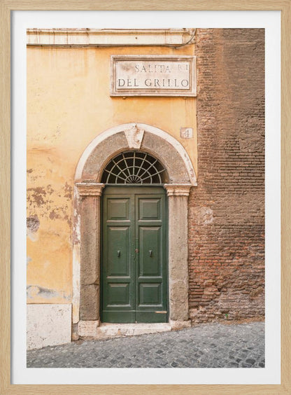 A vertical shot of a rustic European street scene featuring a dark green arched wooden door set into a stone frame. The building's facade is a mix of peeling yellow ochre plaster and exposed red brick. Above the door is a marble plaque that reads 'SALITA RI DEL GRILLO', and the ground is paved with grey cobblestones. Wall Art