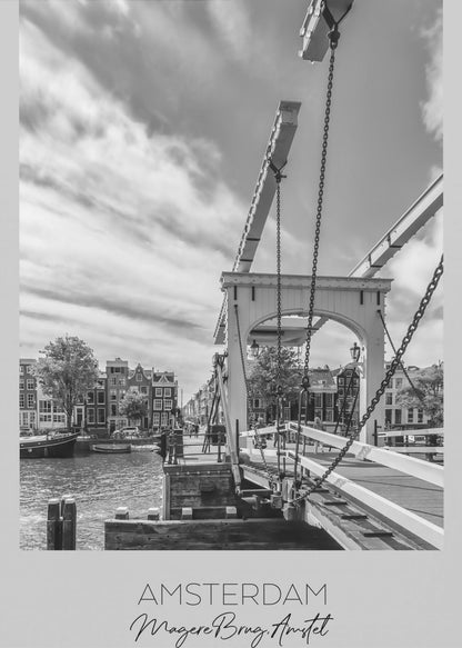 A black and white travel poster of Amsterdam, featuring a side view of the Magere Brug, a traditional wooden drawbridge, over the Amstel river. Classic canal houses line the waterway in the background under a dramatic, cloudy sky. The image is in a black frame and has text at the bottom that reads 'AMSTERDAM', 'Magere Brug, Amstel', and 'NEDERLAND'. Artwork