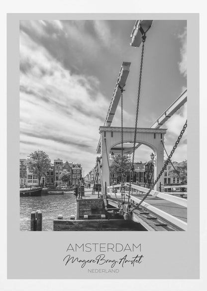 A black and white travel poster of Amsterdam, featuring a side view of the Magere Brug, a traditional wooden drawbridge, over the Amstel river. Classic canal houses line the waterway in the background under a dramatic, cloudy sky. The image is in a black frame and has text at the bottom that reads 'AMSTERDAM', 'Magere Brug, Amstel', and 'NEDERLAND'. Artwork