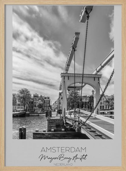 A black and white travel poster of Amsterdam, featuring a side view of the Magere Brug, a traditional wooden drawbridge, over the Amstel river. Classic canal houses line the waterway in the background under a dramatic, cloudy sky. The image is in a black frame and has text at the bottom that reads 'AMSTERDAM', 'Magere Brug, Amstel', and 'NEDERLAND'. Artwork