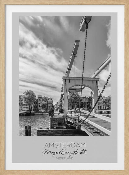 A black and white travel poster of Amsterdam, featuring a side view of the Magere Brug, a traditional wooden drawbridge, over the Amstel river. Classic canal houses line the waterway in the background under a dramatic, cloudy sky. The image is in a black frame and has text at the bottom that reads 'AMSTERDAM', 'Magere Brug, Amstel', and 'NEDERLAND'. Artwork