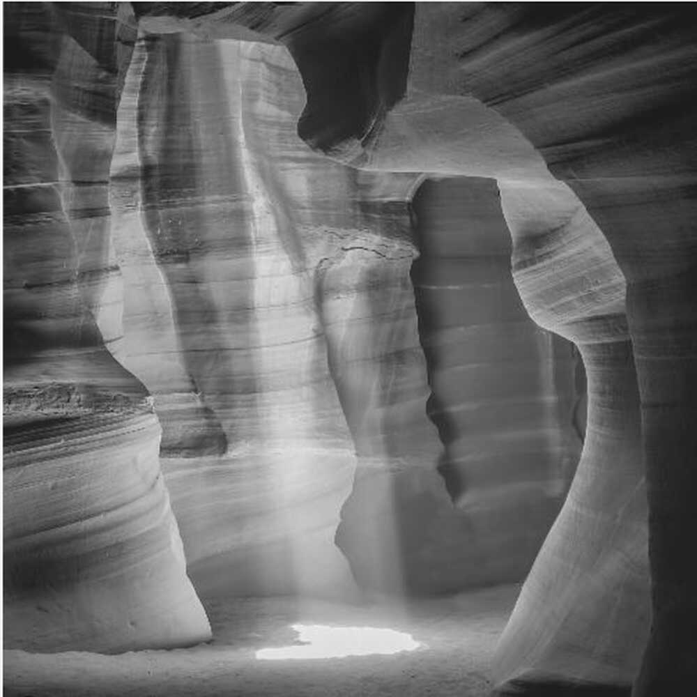 A dramatic black and white photo of a sunbeam lighting the sandy floor of a smooth, curved slot canyon.