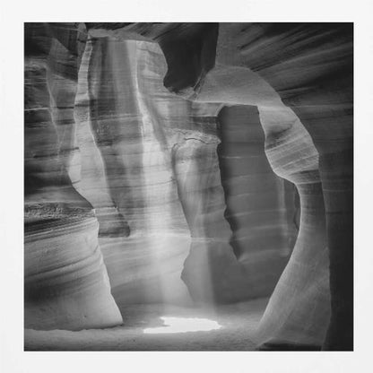 A dramatic black and white photo of a sunbeam lighting the sandy floor of a smooth, curved slot canyon.
