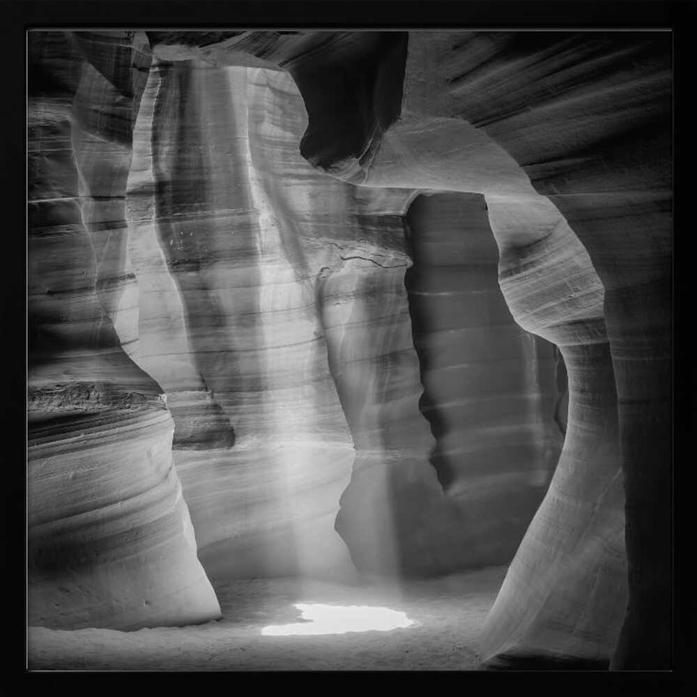 A dramatic black and white photo of a sunbeam lighting the sandy floor of a smooth, curved slot canyon.
