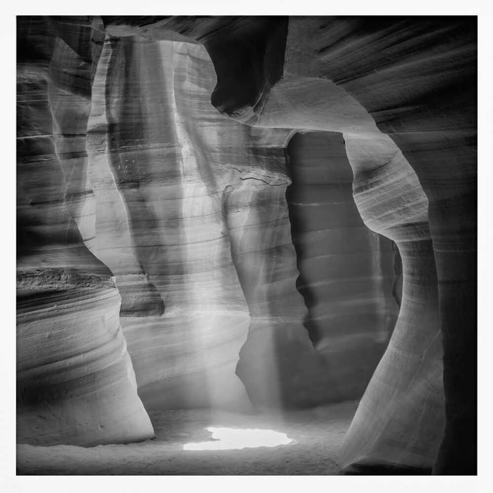 A dramatic black and white photo of a sunbeam lighting the sandy floor of a smooth, curved slot canyon.