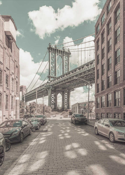 A vertical photograph of the Manhattan Bridge from a cobblestone street in DUMBO, Brooklyn, framed by tall brick buildings. The image has a vintage aesthetic with a muted teal sky and sepia-toned buildings and streets. Print