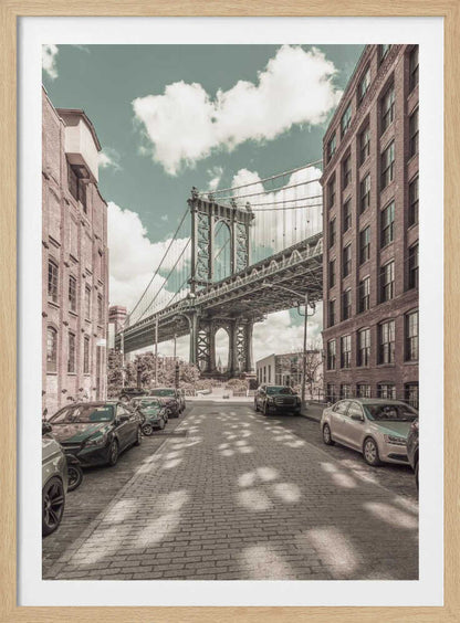 A vertical photograph of the Manhattan Bridge from a cobblestone street in DUMBO, Brooklyn, framed by tall brick buildings. The image has a vintage aesthetic with a muted teal sky and sepia-toned buildings and streets. Print
