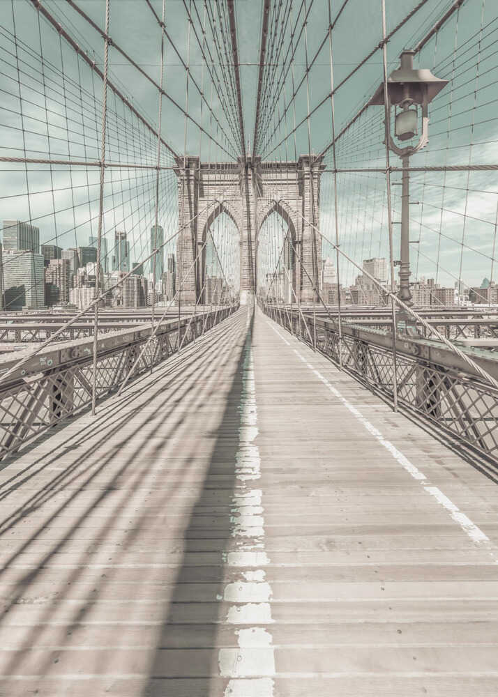 A vintage-style photograph looking down the center of the Brooklyn Bridge's wooden pedestrian walkway towards its iconic stone arches, with the New York City skyline visible in the background under a pale teal sky. The bridge's intricate network of suspension cables creates a strong sense of vanishing point perspective. Decor
