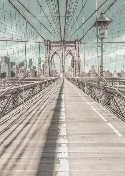 A vintage-style photograph looking down the center of the Brooklyn Bridge's wooden pedestrian walkway towards its iconic stone arches, with the New York City skyline visible in the background under a pale teal sky. The bridge's intricate network of suspension cables creates a strong sense of vanishing point perspective. Decor