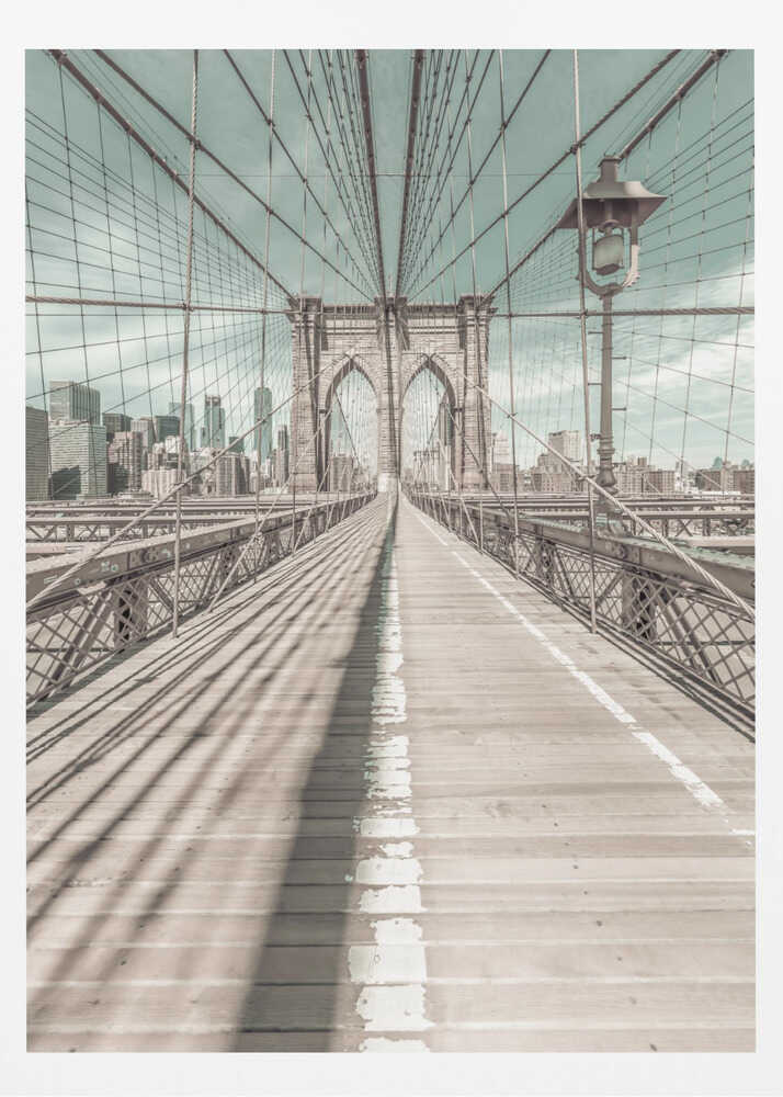 A vintage-style photograph looking down the center of the Brooklyn Bridge's wooden pedestrian walkway towards its iconic stone arches, with the New York City skyline visible in the background under a pale teal sky. The bridge's intricate network of suspension cables creates a strong sense of vanishing point perspective. Decor