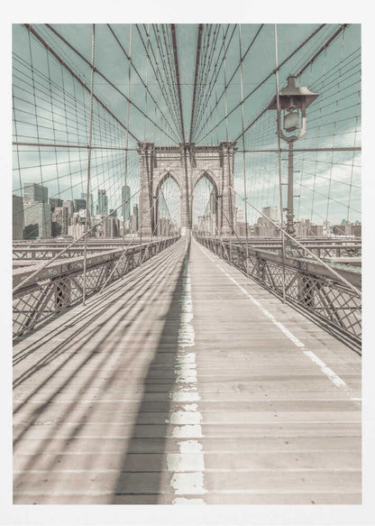 A vintage-style photograph looking down the center of the Brooklyn Bridge's wooden pedestrian walkway towards its iconic stone arches, with the New York City skyline visible in the background under a pale teal sky. The bridge's intricate network of suspension cables creates a strong sense of vanishing point perspective. Decor