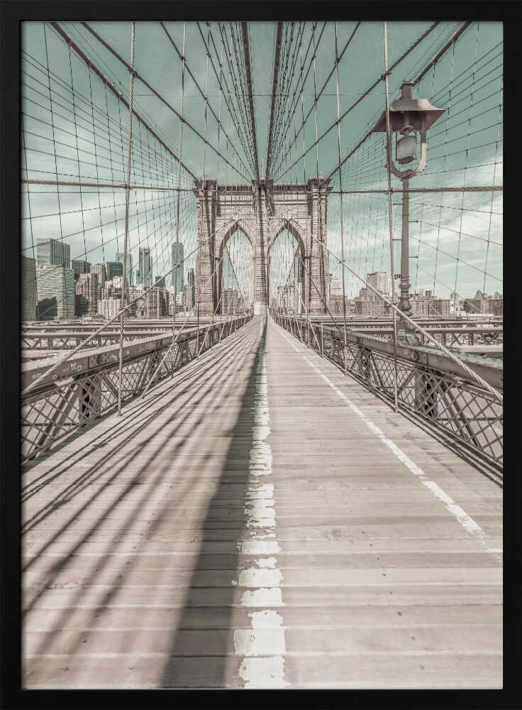 A vintage-style photograph looking down the center of the Brooklyn Bridge's wooden pedestrian walkway towards its iconic stone arches, with the New York City skyline visible in the background under a pale teal sky. The bridge's intricate network of suspension cables creates a strong sense of vanishing point perspective. Decor