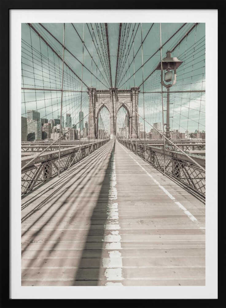 A vintage-style photograph looking down the center of the Brooklyn Bridge's wooden pedestrian walkway towards its iconic stone arches, with the New York City skyline visible in the background under a pale teal sky. The bridge's intricate network of suspension cables creates a strong sense of vanishing point perspective. Decor