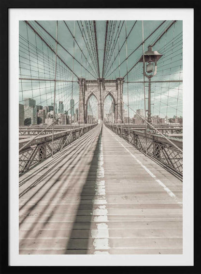 A vintage-style photograph looking down the center of the Brooklyn Bridge's wooden pedestrian walkway towards its iconic stone arches, with the New York City skyline visible in the background under a pale teal sky. The bridge's intricate network of suspension cables creates a strong sense of vanishing point perspective. Decor