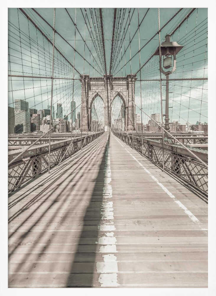 A vintage-style photograph looking down the center of the Brooklyn Bridge's wooden pedestrian walkway towards its iconic stone arches, with the New York City skyline visible in the background under a pale teal sky. The bridge's intricate network of suspension cables creates a strong sense of vanishing point perspective. Decor