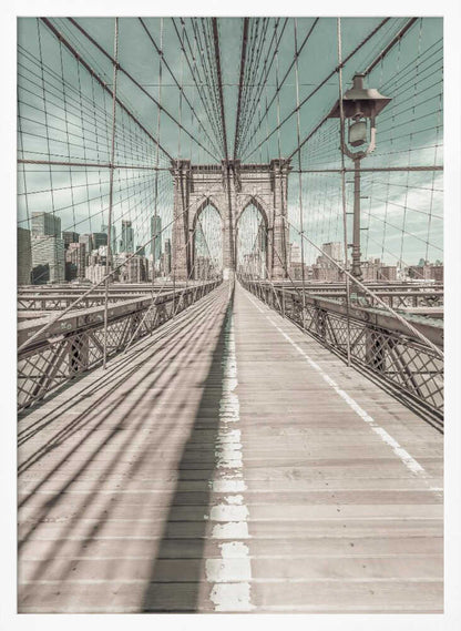 A vintage-style photograph looking down the center of the Brooklyn Bridge's wooden pedestrian walkway towards its iconic stone arches, with the New York City skyline visible in the background under a pale teal sky. The bridge's intricate network of suspension cables creates a strong sense of vanishing point perspective. Decor