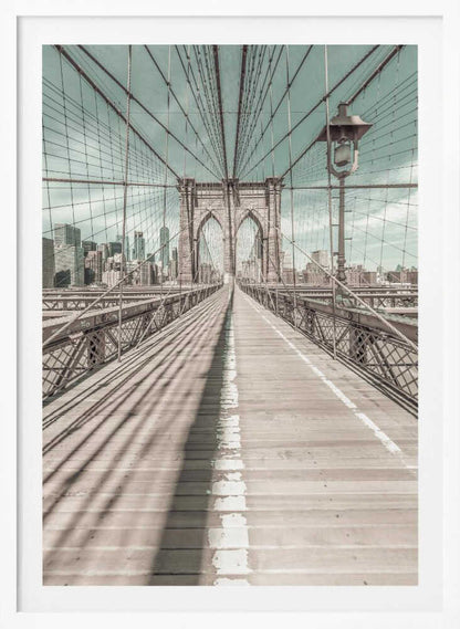 A vintage-style photograph looking down the center of the Brooklyn Bridge's wooden pedestrian walkway towards its iconic stone arches, with the New York City skyline visible in the background under a pale teal sky. The bridge's intricate network of suspension cables creates a strong sense of vanishing point perspective. Decor