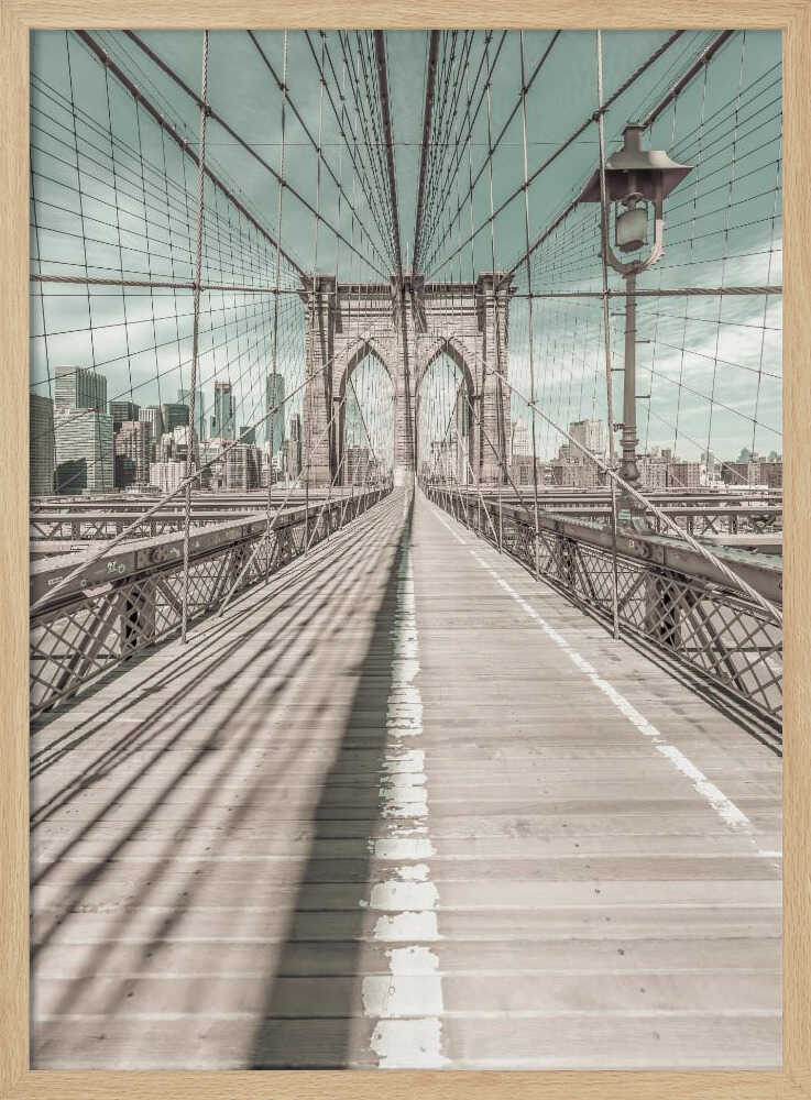 A vintage-style photograph looking down the center of the Brooklyn Bridge's wooden pedestrian walkway towards its iconic stone arches, with the New York City skyline visible in the background under a pale teal sky. The bridge's intricate network of suspension cables creates a strong sense of vanishing point perspective. Decor