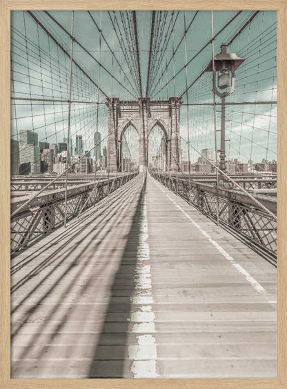A vintage-style photograph looking down the center of the Brooklyn Bridge's wooden pedestrian walkway towards its iconic stone arches, with the New York City skyline visible in the background under a pale teal sky. The bridge's intricate network of suspension cables creates a strong sense of vanishing point perspective. Decor