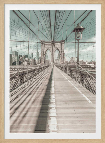 A vintage-style photograph looking down the center of the Brooklyn Bridge's wooden pedestrian walkway towards its iconic stone arches, with the New York City skyline visible in the background under a pale teal sky. The bridge's intricate network of suspension cables creates a strong sense of vanishing point perspective. Decor