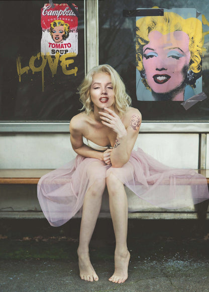 A blonde woman styled like Marilyn Monroe sits on a bench, wearing a pink tulle skirt. Behind her, a dark window features an Andy Warhol-style poster of Monroe, a Campbell's soup can parody, and the word 'LOVE' in gold graffiti. Artwork