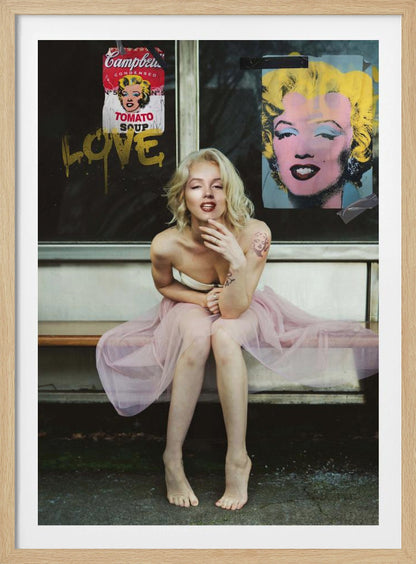 A blonde woman styled like Marilyn Monroe sits on a bench, wearing a pink tulle skirt. Behind her, a dark window features an Andy Warhol-style poster of Monroe, a Campbell's soup can parody, and the word 'LOVE' in gold graffiti. Artwork