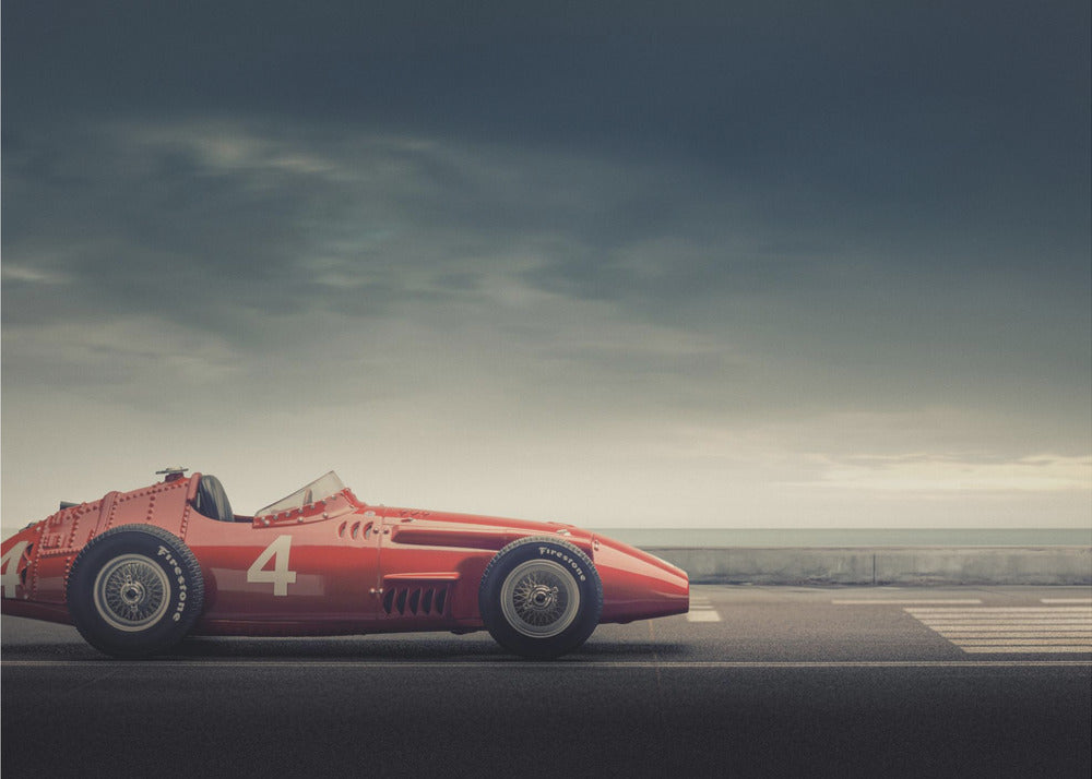 A side profile of a vintage red single-seater race car with the number 4 painted on it, parked on a seaside road under a dramatic, cloudy sky. The image has a muted, nostalgic color palette and is shown within a silver frame. Decor