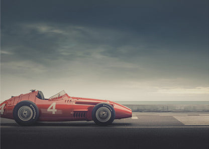 A side profile of a vintage red single-seater race car with the number 4 painted on it, parked on a seaside road under a dramatic, cloudy sky. The image has a muted, nostalgic color palette and is shown within a silver frame. Decor