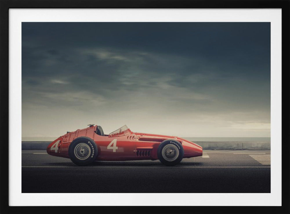A side profile of a vintage red single-seater race car with the number 4 painted on it, parked on a seaside road under a dramatic, cloudy sky. The image has a muted, nostalgic color palette and is shown within a silver frame. Decor