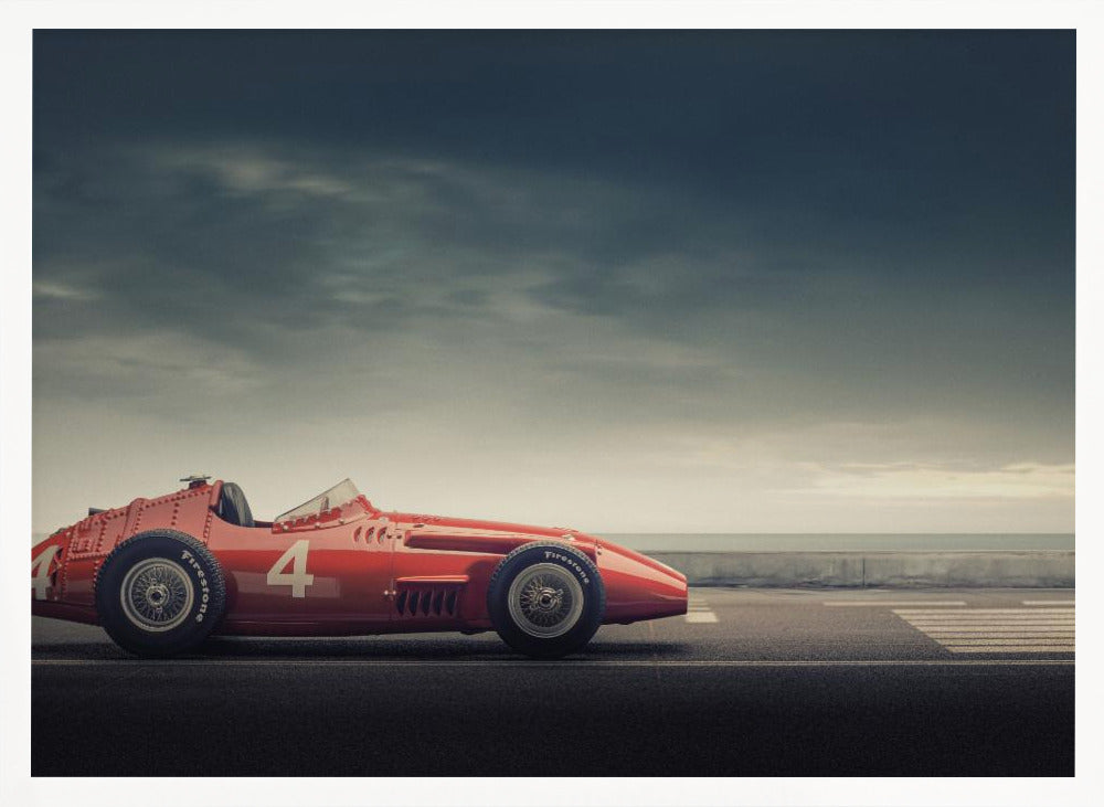 A side profile of a vintage red single-seater race car with the number 4 painted on it, parked on a seaside road under a dramatic, cloudy sky. The image has a muted, nostalgic color palette and is shown within a silver frame. Decor