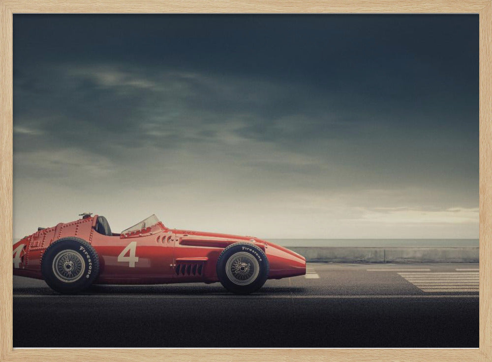 A side profile of a vintage red single-seater race car with the number 4 painted on it, parked on a seaside road under a dramatic, cloudy sky. The image has a muted, nostalgic color palette and is shown within a silver frame. Decor