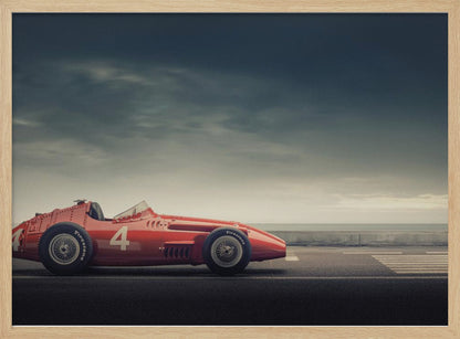 A side profile of a vintage red single-seater race car with the number 4 painted on it, parked on a seaside road under a dramatic, cloudy sky. The image has a muted, nostalgic color palette and is shown within a silver frame. Decor