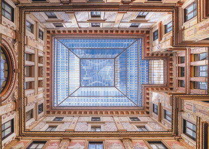 A worm's-eye view looking straight up from the center of an ornate, multi-story courtyard. The detailed walls create a symmetrical frame around a large glass skylight, which reflects a bright blue sky with fluffy white clouds. Print