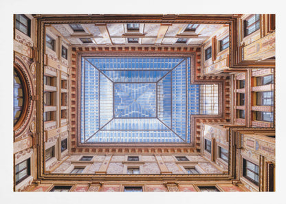 A worm's-eye view looking straight up from the center of an ornate, multi-story courtyard. The detailed walls create a symmetrical frame around a large glass skylight, which reflects a bright blue sky with fluffy white clouds. Print
