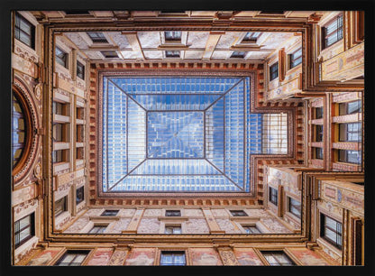 A worm's-eye view looking straight up from the center of an ornate, multi-story courtyard. The detailed walls create a symmetrical frame around a large glass skylight, which reflects a bright blue sky with fluffy white clouds. Print