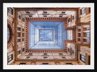 A worm's-eye view looking straight up from the center of an ornate, multi-story courtyard. The detailed walls create a symmetrical frame around a large glass skylight, which reflects a bright blue sky with fluffy white clouds. Print