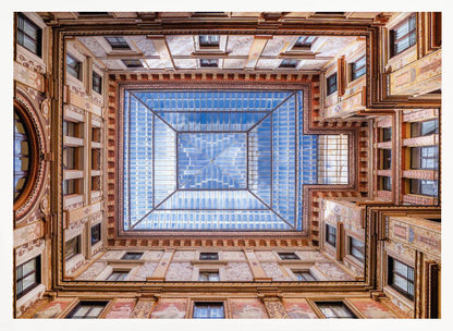 A worm's-eye view looking straight up from the center of an ornate, multi-story courtyard. The detailed walls create a symmetrical frame around a large glass skylight, which reflects a bright blue sky with fluffy white clouds. Print