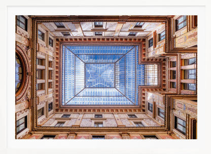 A worm's-eye view looking straight up from the center of an ornate, multi-story courtyard. The detailed walls create a symmetrical frame around a large glass skylight, which reflects a bright blue sky with fluffy white clouds. Print