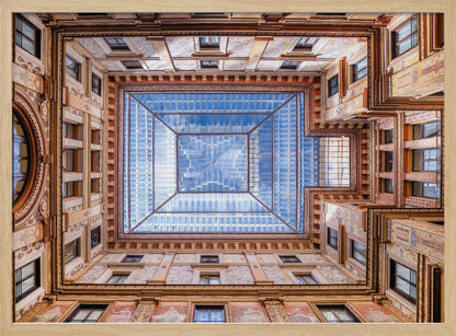 A worm's-eye view looking straight up from the center of an ornate, multi-story courtyard. The detailed walls create a symmetrical frame around a large glass skylight, which reflects a bright blue sky with fluffy white clouds. Print