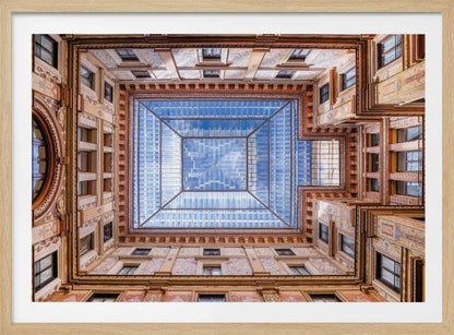 A worm's-eye view looking straight up from the center of an ornate, multi-story courtyard. The detailed walls create a symmetrical frame around a large glass skylight, which reflects a bright blue sky with fluffy white clouds. Print