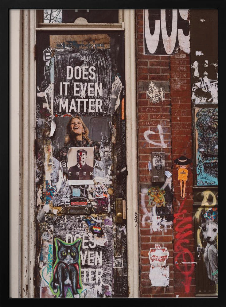 A close-up of a weathered dark door and an adjacent brick wall, both covered in a dense collage of street art, including peeling posters, stickers, and graffiti. A prominent poster on the door asks 'DOES IT EVEN MATTER' in bold white letters. Wall Art