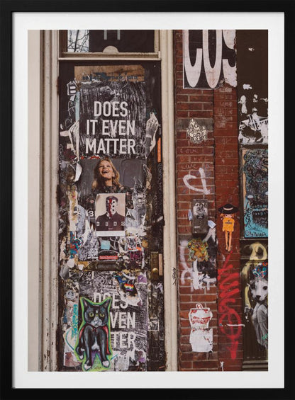A close-up of a weathered dark door and an adjacent brick wall, both covered in a dense collage of street art, including peeling posters, stickers, and graffiti. A prominent poster on the door asks 'DOES IT EVEN MATTER' in bold white letters. Wall Art