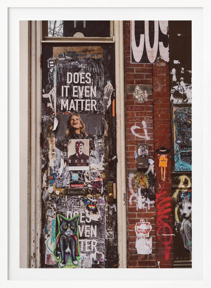 A close-up of a weathered dark door and an adjacent brick wall, both covered in a dense collage of street art, including peeling posters, stickers, and graffiti. A prominent poster on the door asks 'DOES IT EVEN MATTER' in bold white letters. Wall Art